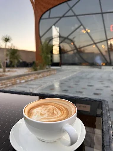 Latte art in a white cup on a table, with a modern restaurant exterior and sunset in the background.